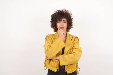 Düşünceli gülümseyen kızın portresi elini çenesinin altında tutuyor, doğrudan kameraya bakıyor, ilgiyle karışık bir şey dinliyor, sıradan giyiniyor, gri duvara karşı poz veriyor. Gençlik kavramı.