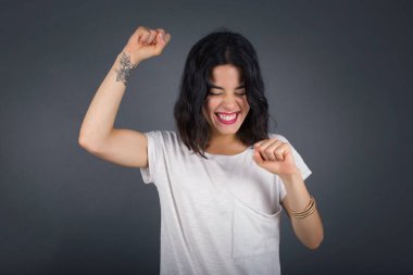 Çekici beyaz kadın zaferi kutluyor yumruklarıyla gökyüzünü yumrukluyor ve gri bir stüdyo arka planında ışıltılı bir gülümsemeyle fotokopi çekiyor. 