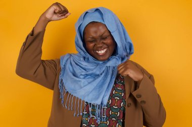 Çekici genç Afrikalı Amerikalı Müslüman kadın zaferi kutluyor yumruklarıyla havaya yumruk atıyor ve sarı renkli bir tebessümle arka planda fotokopi çekiyordu.