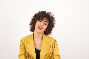 Geniş tebessümlü güzel esmer kadın, beyaz dişlerini gösteriyor, günlük tişört giyiyor, gri stüdyo duvarının başında dikiliyor izin gününün keyfini çıkarıyor. Kadın içeride duruyor..