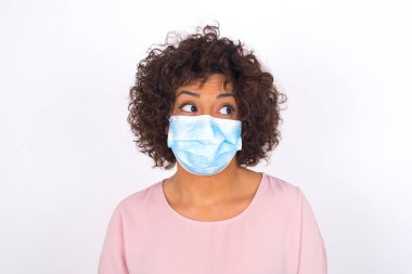 Yüz maskesi takan ve sıradan giyinen bir kadın beyaz stüdyo arka planında izole edilmiş bir şekilde yüzünü çeviriyor.