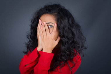 Güzel, genç, kıvırcık saçlı, kırmızı gömlekli, elleriyle yüzünü örten ve parmaklarının arasından dışarı bakan bir kadın..