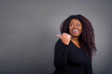 Omg konsepti. Şaşırmış bir ifadeyle afro-amerikan kadın, gözlerini ve ağzını iyice açıyor, baş parmağıyla işaret ediyor, gri arka planda garip bir şey gösteriyor. Reklam kavramı.