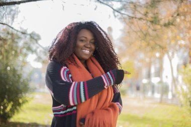 Güz parkını gösteren uzun saçlı, renkli kazaklı güzel Afro-Amerikan kadın.  