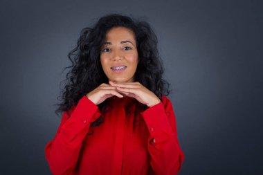 Kıvırcık saçlı, kırmızı gömlekli, gri arka planda poz veren ve çenesine elleriyle dokunan sevimli esmer kadın.