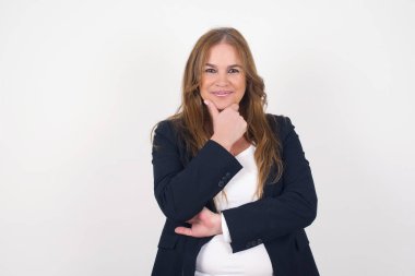 Düşünceli orta yaşlı beyaz kadın portresi elini çenesinin altında tutuyor, doğrudan kameraya bakıyor, ilgi çekici bir şey dinliyor, sıradan giyiniyor, gri duvara karşı poz veriyor.. 