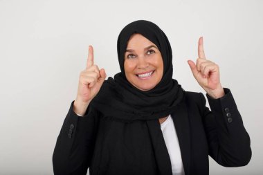 Siyah tesettürlü başarılı müslüman kadının yatay görüntüsü, her iki işaret parmağını havaya kaldırarak, boş kopya uzay duvarında şok edici bir şeyi işaret ediyor..