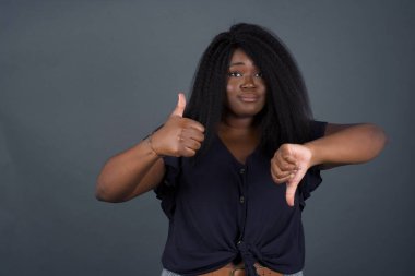 Güzel Afro-Amerikan kadın iyiye-kötüye işaret veriyor. Hoşnutsuz ve etkilenmemiş, sıradan kıyafetler giyiyor ve gri duvarın önünde dikiliyor..