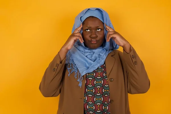 Dreamy African American muslim woman with thoughtful expression, looks away, keeps hand near face, thinks about something pleasant, poses against yellow wall.