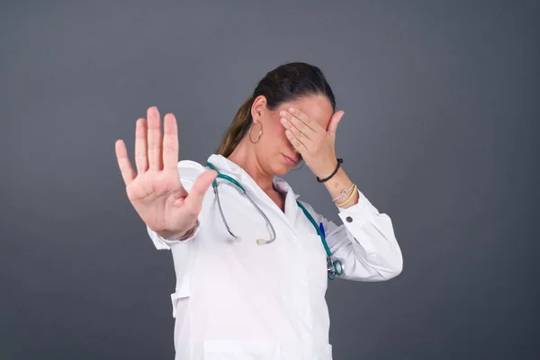 People, body language. Young European doctor woman covers eyes with palm and doing stop gesture, tries to hide from everybody. Don\'t look at me, I don\'t want to see, feels ashamed or scared.