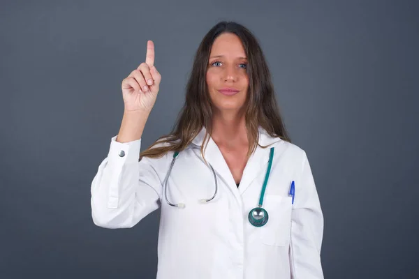 身穿医疗制服的年轻的高加索女医生 站在灰墙旁边 露出并用第一指尖指尖 面带微笑 自信而快乐 — 图库照片