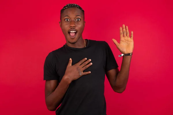 I swear, promise you not regret. Portrait of happy, sincere young African American man wearing black t-shirt over red background raising one arm and hold hand on heart as give oath, pledge, telling truth, want you to believe