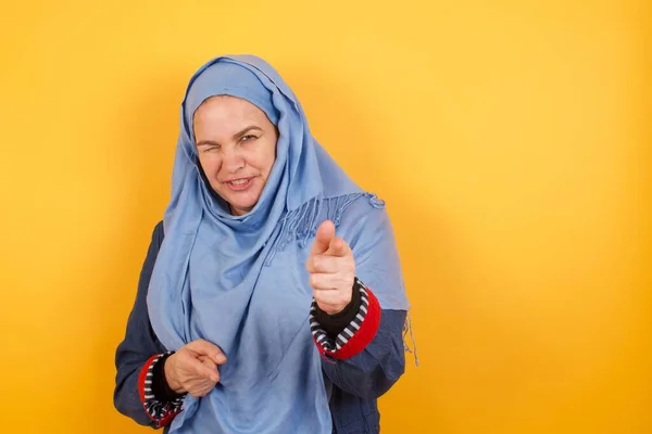ヒジャーブウィンキングの喜びとカリスマ的な見栄えの良い中年のイスラム教徒の女性と喜んで壁にポーズするカメラで指ピストルを指差します — ストック写真