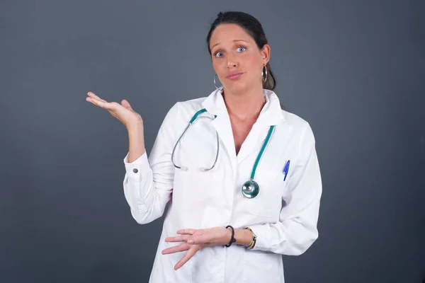 一位美丽的欧洲医生女士被灰色背景隔离 双手都伸出来 露出奇怪的表情 我不知道这是什么 站在灰色的背景下 广告概念 — 图库照片