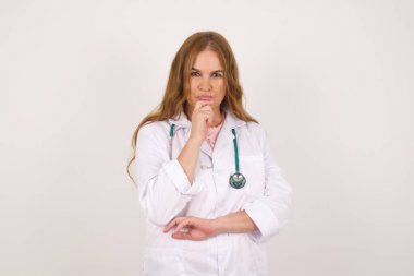 Medikal üniforma giyen düşünceli, olgun, beyaz kadın doktor elini çenesinin altında tutuyor, düşünceli bir şekilde yukarı bakıyor, tıbbi üniforma giyiyor, beyaz arka plan üzerinde poz veriyor. 