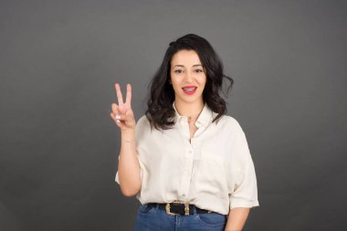 Güzel esmer kadın, sıradan giyinmiş, zafer jestleriyle. Gri arkaplanda duruyor. 