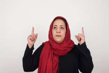 Başarılı, dost canlısı, parlak makyajlı, parlak saçlı bir kadının işaret parmağıyla işaret ederek boş duvarda şok edici bir şey gösterdiği yatay bir görüntü. Stüdyoda poz veren başörtüsü takan orta yaşlı bir Arap kadın.