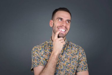 Hayalperest bir erkek, düşünceli bir ifadeyle, kameraya bakıyor, elini yüzüne yakın tutuyor, parmağını ısırıyor, hoş bir şey düşünüyor, gri duvara karşı poz veriyor..