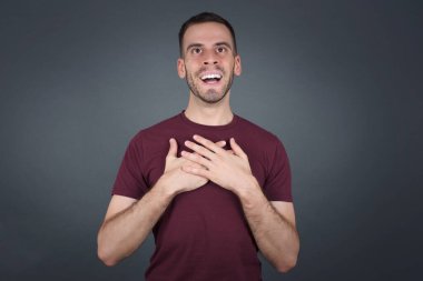 Mutlu adam ayakta ve gülümsüyor. Gri stüdyo arka planında tek başına. Elleri göğsüne yakın kalbi var. Genç duygusal adam. İnsani duygular, yüz ifadesi kavramı. Ön görünüm.