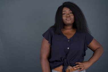 Neşeli Afrikalı Amerikalı kadının stüdyo çekimi elini kalçasında tutuyor, geniş bir şekilde gülümsüyor, arkadaşlarının doğum gününde hazırladığı sürprizi bekliyor, gri arka planda izole edilmiş..