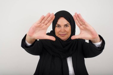 El hareketli parmak çerçevesi. Siyah tesettürlü, gülümseyen Müslüman bir kadının portresi. Kameraya bakıyor ve kapalı alanda dururken el kol hareketi yapıyor.