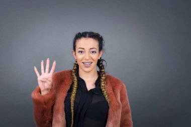 Genç esmer kadın gri duvarın önünde duruyor. Dört numaralı parmaklarıyla işaret ediyor ve gülümsüyor. Kendinden emin ve mutlu..