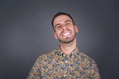 Geniş tebessümlü, beyaz dişleri olan, sıradan tişörtler giyen, gri stüdyo duvarının üzerinde duran yakışıklı bir erkek izinli olduğu için seviniyor. İçerde dikilen adam.