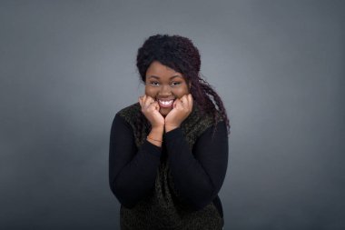 Hayalperest Afro-Amerikan kadını ellerini çenesinin altında tutuyor, mutlu bir ifadeyle bakıyor, dişlek gülüşü var, gri arka planda izole edilmiş.