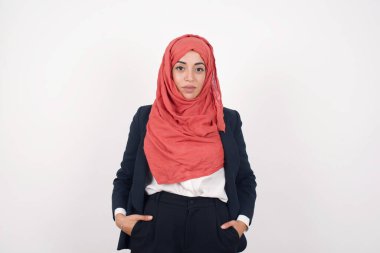 Güzel Müslüman kadın siyah ceket giyer ve izole edilmiş arka planda tesettür takar. Elleri ceplerinde kameraya bakarak gülümserler. Pozitif kişi.