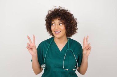 Tıp üniforması giyen güzel bir Arap doktorun yalnız fotoğrafı iki eliyle barış ya da zafer işareti yapar, rahat kıyafetler giyer, havalı hisseder, dişlek bir gülümsemesi vardır, arka planda izole olur. İnsanlar ve vücut dili.