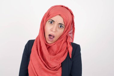 Etkileyici yüz ifadeleri. Şok olmuş güzel Müslüman kadın siyah ceket giyiyor ve başörtüsü düşük çenesini tutuyor, çizgili bir süveter giyiyor, boş gri duvarda gördüklerinden sersemlemiş hissediyor.