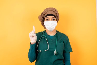 Yeşil üniformalı, genç, Müslüman doktor kadın. Duvara yaslanmış, kendini beğenmiş ve mutlu bir şekilde gülümserken bir numaralı parmaklarıyla işaret ediyor..