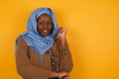 Neşeli, güzel, Afro-Amerikan Müslüman bir kadın resmi. Sarı duvarın karşısında duruyor. Yüzü birbirine yakın. İyi haberleri dinledikten sonra kameraya mutlu bir ifadeyle bakmak. Kendine güvenen kız..