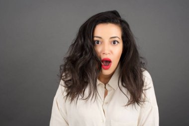 Gayri resmi giyinmiş güzel esmer kadın şaşırdı. Gri arkaplanda duruyor.  