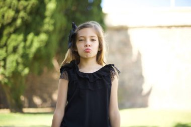 Çekici, açık renk saçlı, siyah elbiseli tatlı kız dudaklarını yuvarlıyor, günlük kıyafetler giyiyor, doğrudan kameraya bakıyor ve memnuniyetini ifade ediyor.