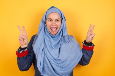 Orta yaşlı, tesettürlü Müslüman bir kadının kapalı bir portresi. Arkaplanda iyimser bir gülümsemeyle izole edilmiş. İki elinle barış ve zafer jesti sergiliyor. V işareti.