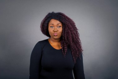 Çekici, açık renk saçlı, sıradan giysili Afrikalı Amerikalı kadın dudakları yuvarlak, günlük kıyafetler giyiyor, doğrudan kameraya bakıyor, memnuniyetini ifade ediyor, gri arka planda izole edilmiş. İnsanlar, vücut dili kavramı.