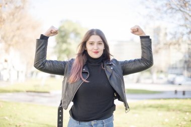 Genç, beyaz tenli, esmer kadın. Bahar parkında deri ceket giyiyor. Kaslarını gösteriyor.
