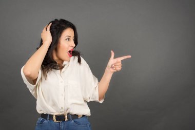 Güzel esmer kadın, sıradan giyinmiş, işaret ediyor. Gri arkaplanda duruyor. 