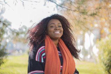 Genç, güzel, gülümseyen, uzun kıvırcık saçlı, Afro-Amerikan kadın. Parkta renkli bir kazak giyiyor.  