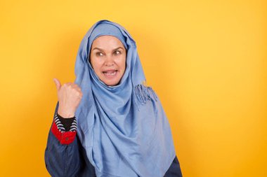 Omg konsepti. Şaşırmış, orta yaşlı, tesettürlü Müslüman bir kadın gözlerini ve ağzını geniş bir şekilde açıyor, başparmağıyla işaret ediyor, arka planda garip bir şey gösteriyor. Reklam kavramı.