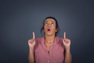 Gri arka planda zarif bir kadının portresi. Şaşırmış, şaşırmış görünüyor ve parmaklarıyla garip bir şey gösteriyor..