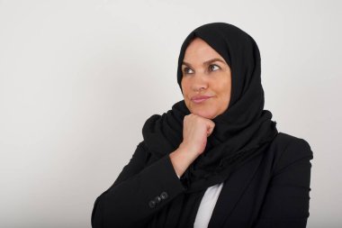 Siyah tesettürlü şık müslüman kadın portresi. Çenesinin altında elleri var ve şüpheli ve şüpheci bir ifadeyle yana bakıyor. Şüpheli ve şüphe dolu. Arka planda kapalı duruyor..