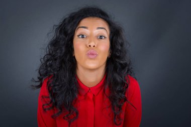 Hoş görünümlü, kıvırcık saçlı, kırmızı tişörtlü esmer bir kadın birini öpmeye giderken dudaklarını tutar..