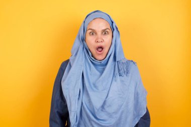 Duygusal olarak çekici, orta yaşlı, tesettürlü, ağzı açık Müslüman bir kadın büyük bir şaşkınlık ve korku içinde, beyaz beton zemine karşı duruyor, kameraya bakıyor.. 