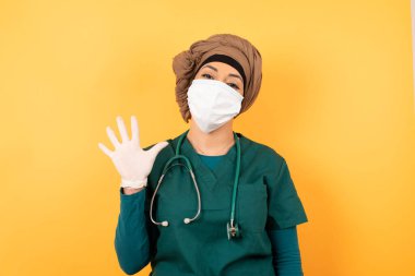 Yeşil üniformalı, genç, güzel, Müslüman doktor kadın duvarın karşısında duruyor ve beş numaralı parmaklarıyla işaret ederken kendinden emin ve mutlu gülümsüyor..