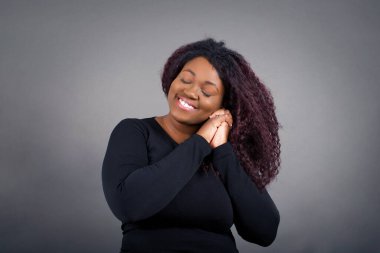 Hayalperest Afro-Amerikan kadını hoş bir ifade takınıyor, gözlerini kapatıyor, ellerini yüzüne çarpıyor, hoş bir şey düşünüyor, gri arka plana karşı poz veriyor..