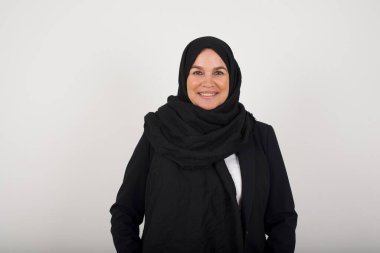 Güven ve iş kavramı. Siyah tesettürlü başarılı Müslüman kadının portresi, gri duvarın önünde el ele tutuşurken kendinden emin bir ifadeyle geniş bir gülümseme sergiliyor..
