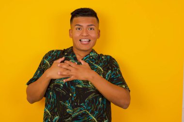 Mutlu İspanyol adam ayakta ve gülümsüyor. Gri stüdyo arka planında tek başına. Göğsüne yakın elleri var. Genç duygusal adam. İnsani duygular, yüz ifadesi kavramı. Ön görünüm.