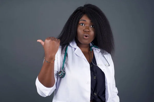 Horizontal shot of pretty young joyful shocked  African American woman doctor points with thumb away, indicates something on blank wall isolated over gray background. Check this out. Ethnicity concept.